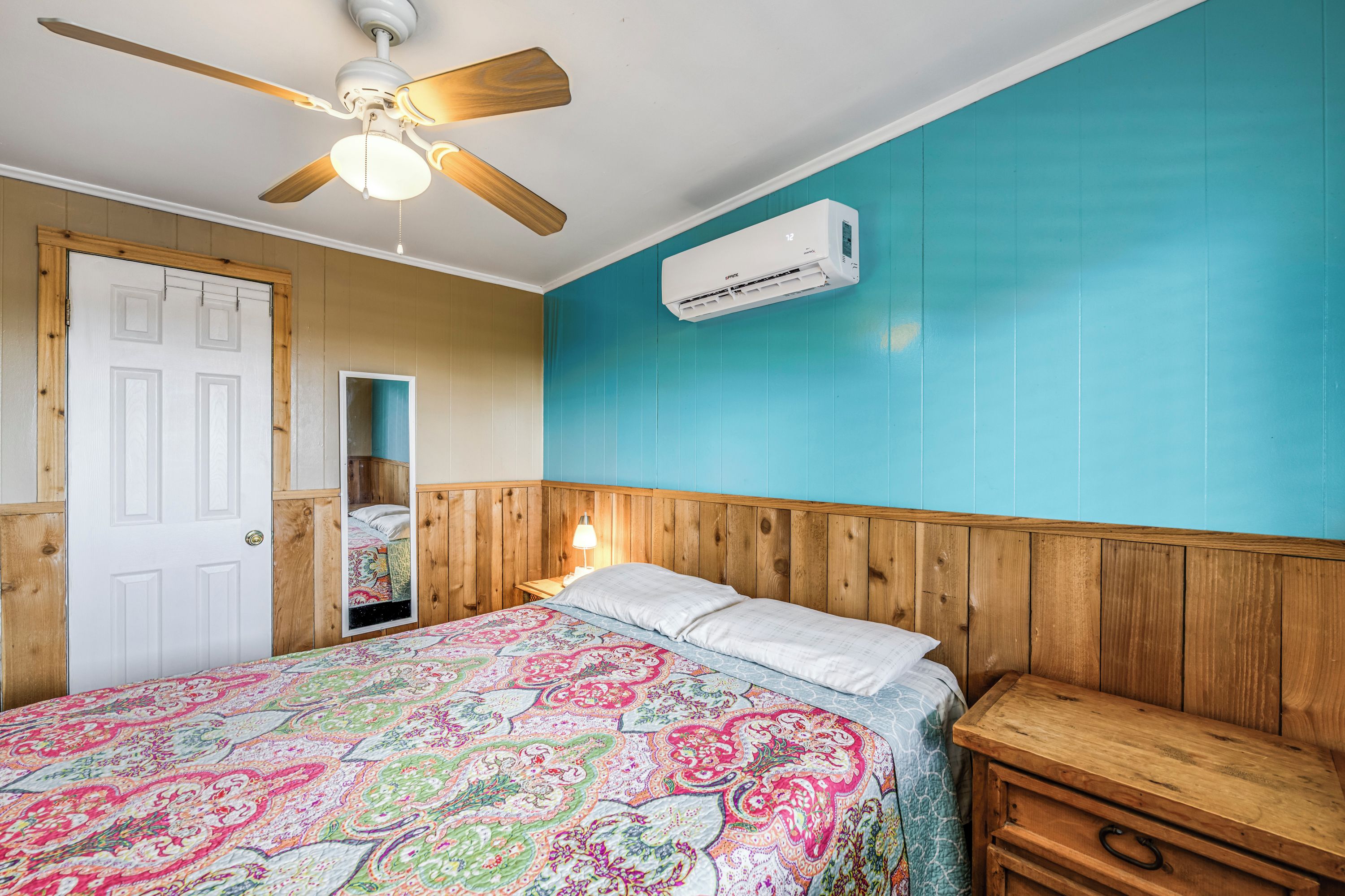 Ein Schlafzimmer mit einem hölzernen Kopfteil, einem Bett mit farbenfroher, gemusterter Bettdecke, einem Deckenventilator und einer Klimaanlage.