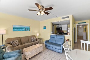 A living room with a brown sofa, a blue armchair, a white table, and a ceiling fan. - BraveWave 1BR King Bed Bay Views Pool & Pier at Laguna Reef (Rockport)