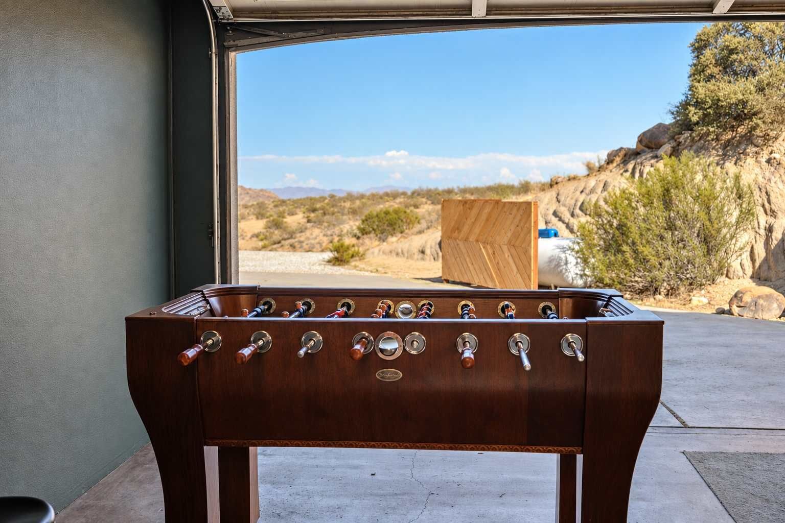 Uma mesa de futebol de madeira com vários controles, posicionada em frente a uma porta de garagem com vista para uma paisagem desértica.