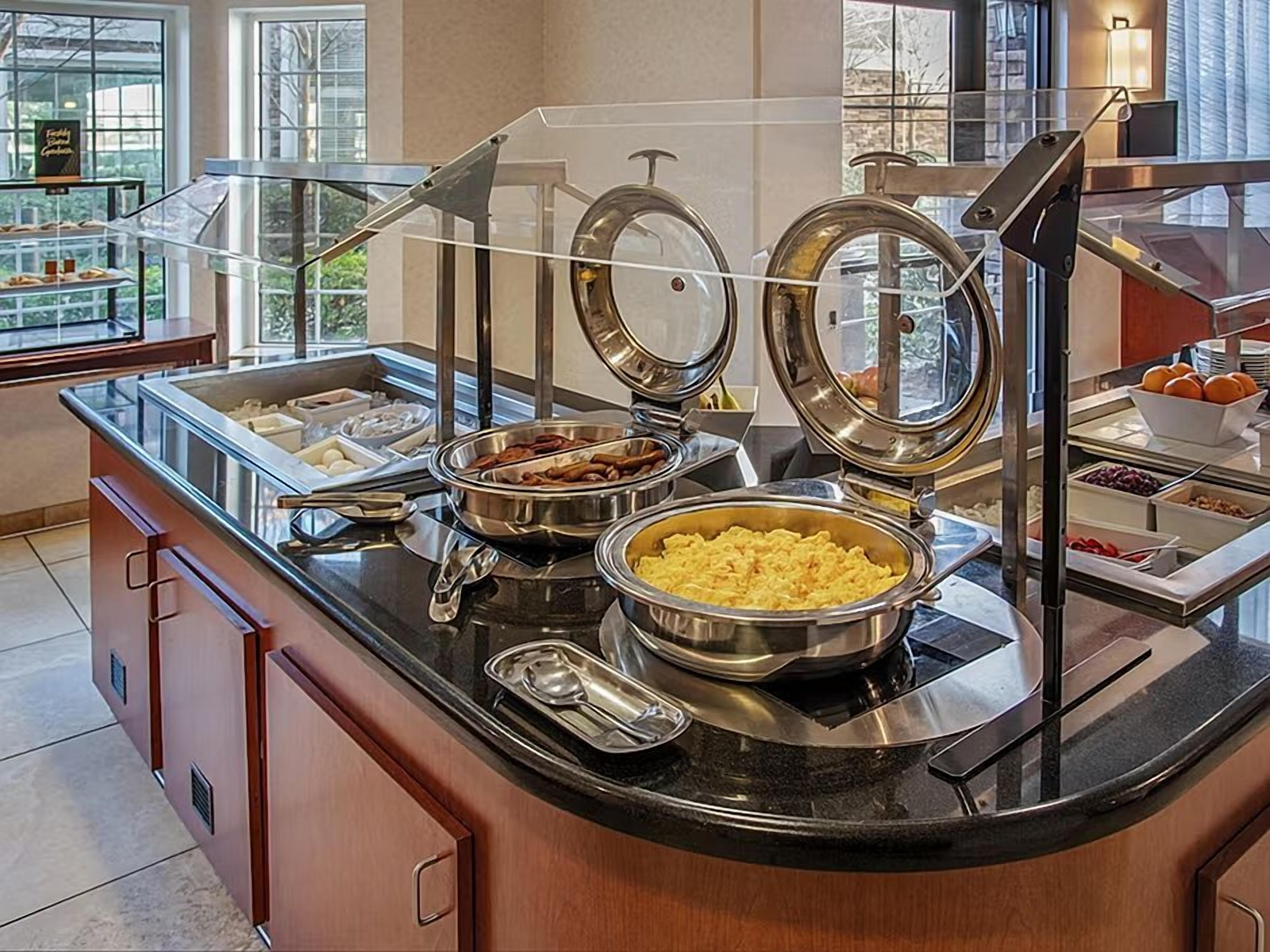 A buffet setup with various food items, including a bowl of scrambled eggs, a container of meat, and a bowl of fruit.