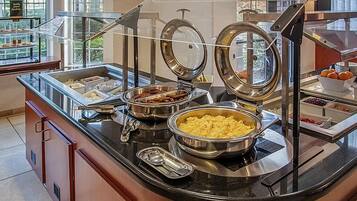 A buffet setup with various food items, including a bowl of scrambled eggs, a container of meat, and a bowl of fruit.