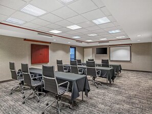 Un salón de conferencias con piso alfombrado, varias mesas con sillas, una pantalla de proyector y una puerta.