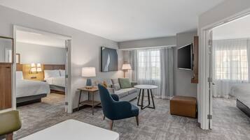 Una habitación de hotel con cama, sofá, sillón, una mesita, televisión y ventana con cortinas.