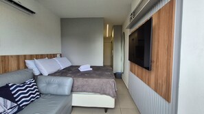 A compact hotel room with a bed, a grey sofa, a TV mounted on the wall, and a wooden headboard. - High Tide - Poolside, apartment 012 by Atlântico Facility & Temporada (Tamandaré)
