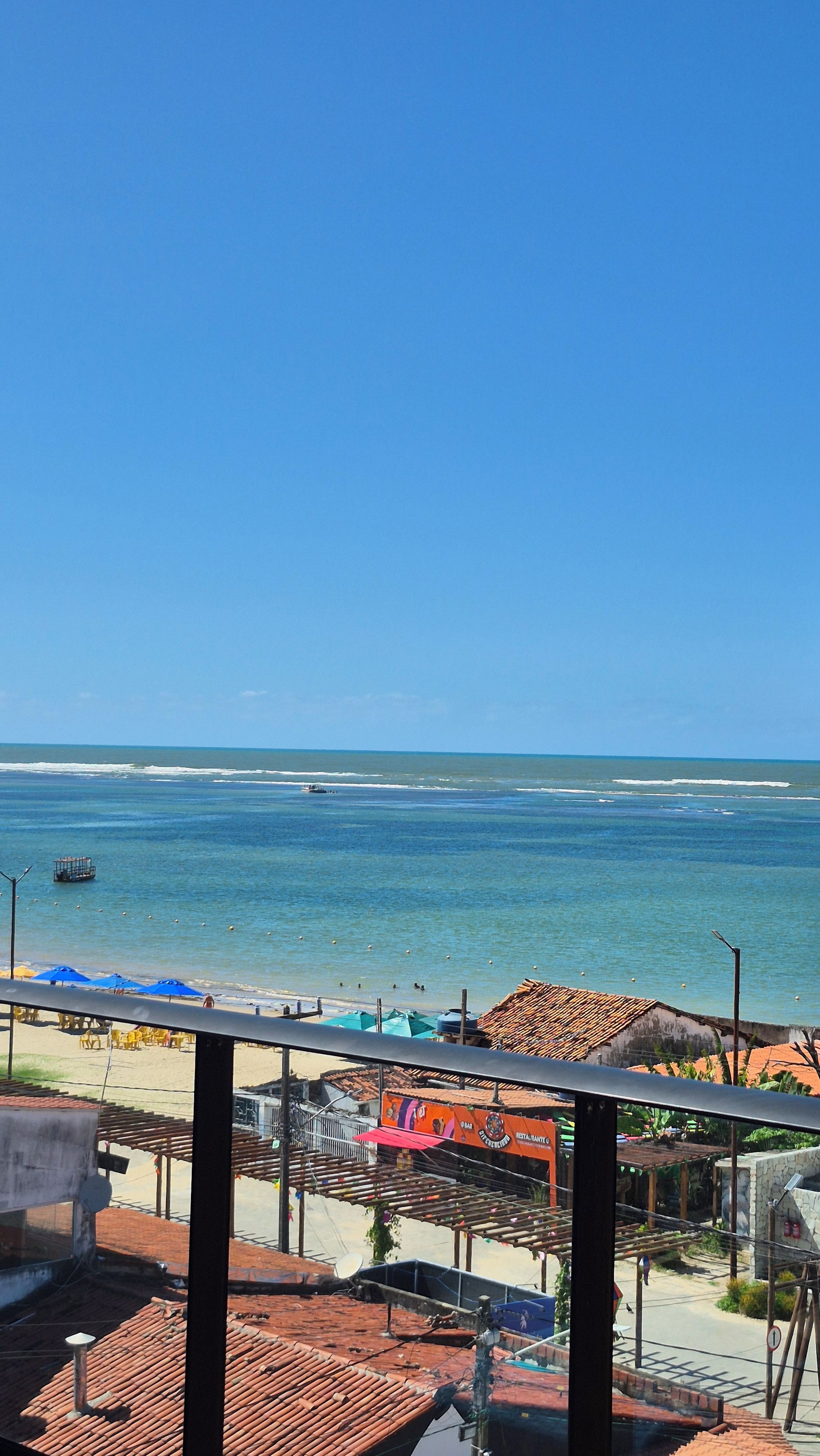 Une vue sur la plage depuis un balcon, avec un ciel dégagé, une plage de sable et quelques parasols.