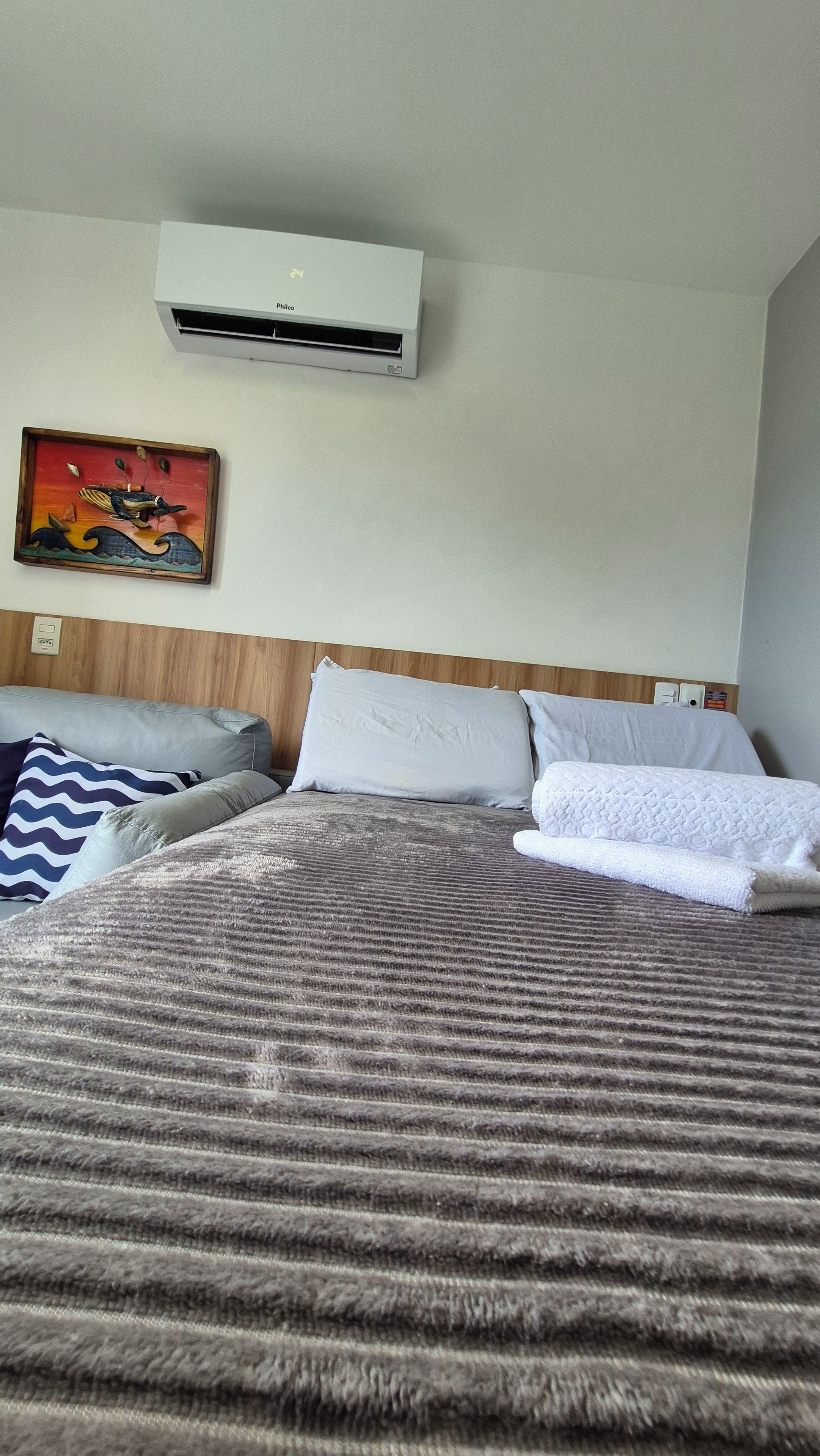 Une chambre d’hôtel comprenant un lit, des oreillers, une tête de lit, un climatiseur fixé au mur et une œuvre d’art encadrée.