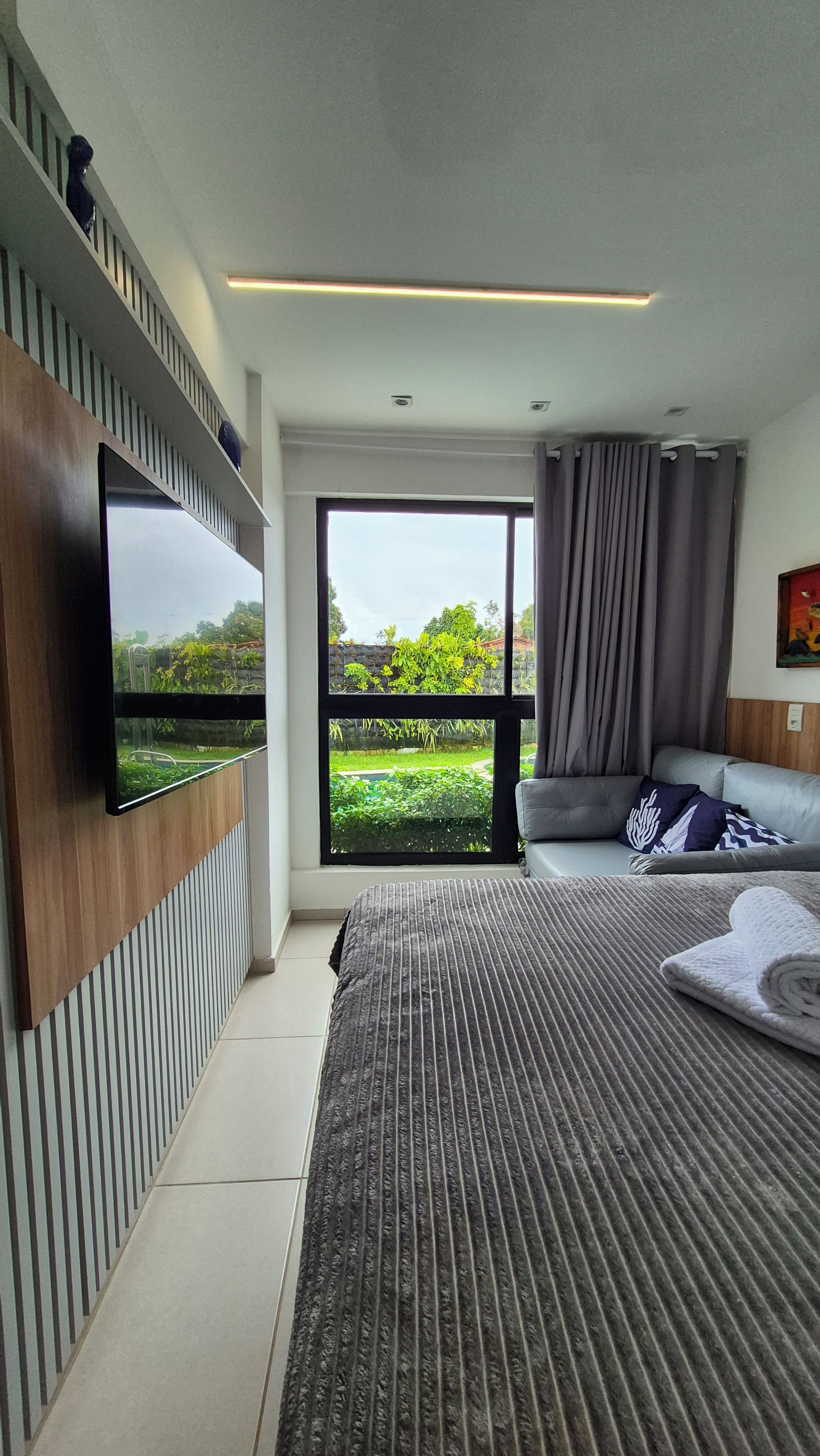 Una camera da letto moderna con un letto grande, una TV a schermo piatto montata a parete, un divano e vista su un giardino attraverso un'ampia finestra.