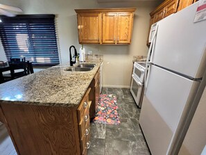 A kitchen with wooden cabinets, a granite countertop, a stainless steel sink, a refrigerator, and a tiled floor. - Tranquil 1-BR condo/St. George/Pools/Gym/near Zion NP & Tuacahn (St. George)