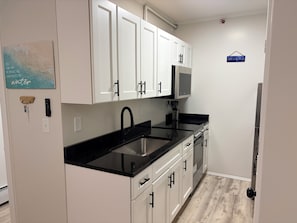 A kitchen with white cabinets, a black countertop, a stainless steel sink, and a microwave. - Steps to Beach & Pier Stylish OOB Studio + Parking (Old Orchard Beach)