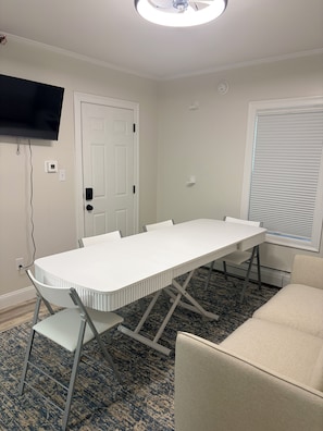 Une salle de conférence avec une table blanche, des chaises, un ventilateur de plafond, un téléviseur fixé au mur et une fenêtre avec des stores.