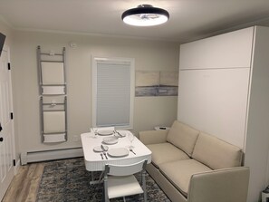 Ein kleiner Wohnraum mit Esstisch für vier Personen, einer Couch, einer Leiter und einem Fenster mit Jalousien.