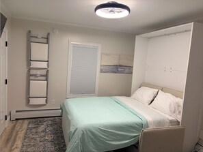 Ein Schlafzimmer mit einem Bett, einem Fenster mit Jalousien, einer Deckenleuchte, einem an der Wand befestigten Regal und einer Tür.