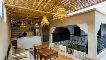 Un área de estar al aire libre con mesas de madera y sillas tejidas, un techo de paja y una cocina en el fondo.