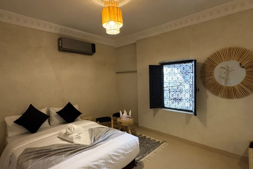 Habitación de hotel con cama, ventana con celosía decorativa, aire acondicionado de pared y un espejo redondo con diseño de sol.