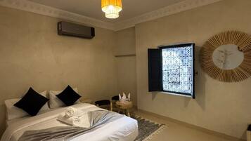 Habitación de hotel con cama, ventana con celosía decorativa, aire acondicionado de pared y un espejo redondo con diseño de sol.