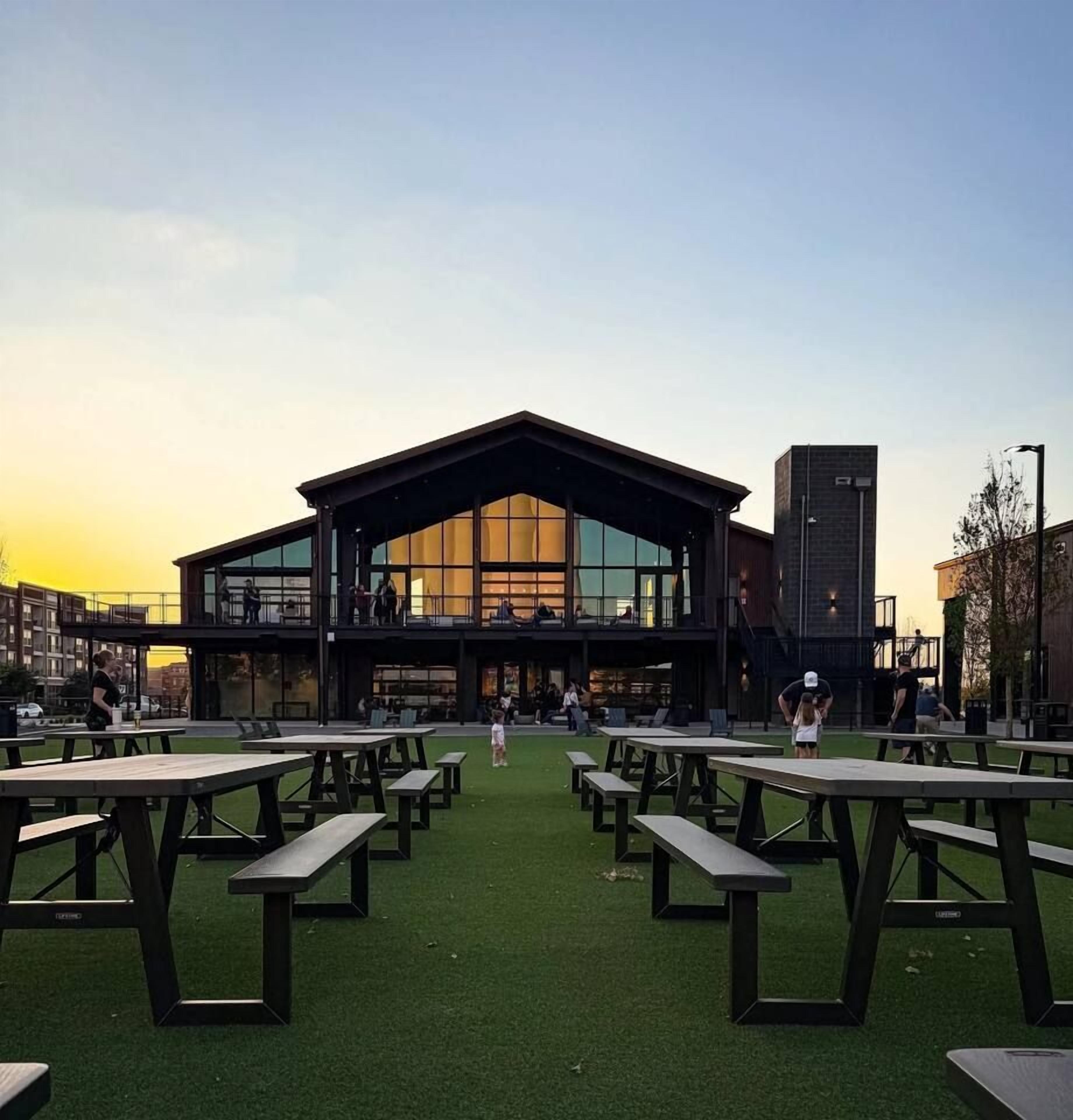 A building with a large glass facade and a flat roof, surrounded by picnic tables on a green lawn.