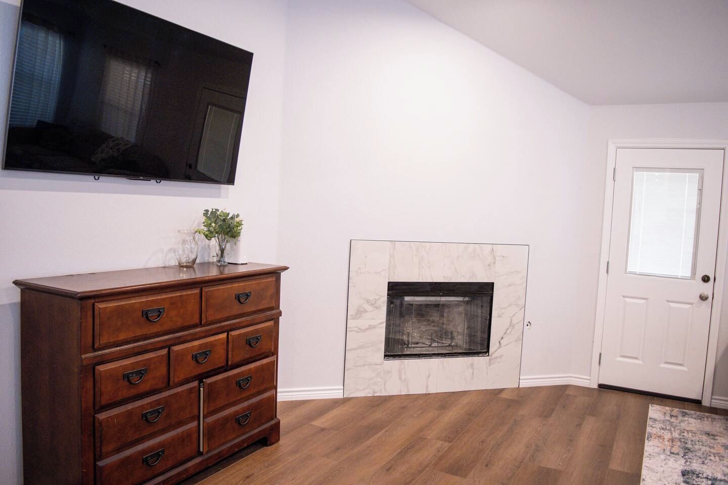 A room with a wooden dresser, a flat-screen TV mounted on the wall, a fireplace with a marble surround, and a white door.