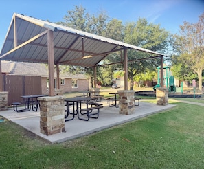 A covered picnic area with tables and benches, a playground with a slide, and a brick building in the background. - Near Lake Retreat | Hot Tub & Boat Slip, High Wifi (Little Elm)