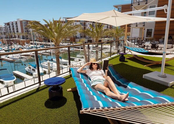 Une personne se détend dans un hamac au bord d’une piscine, avec vue sur des bateaux et des bâtiments modernes.