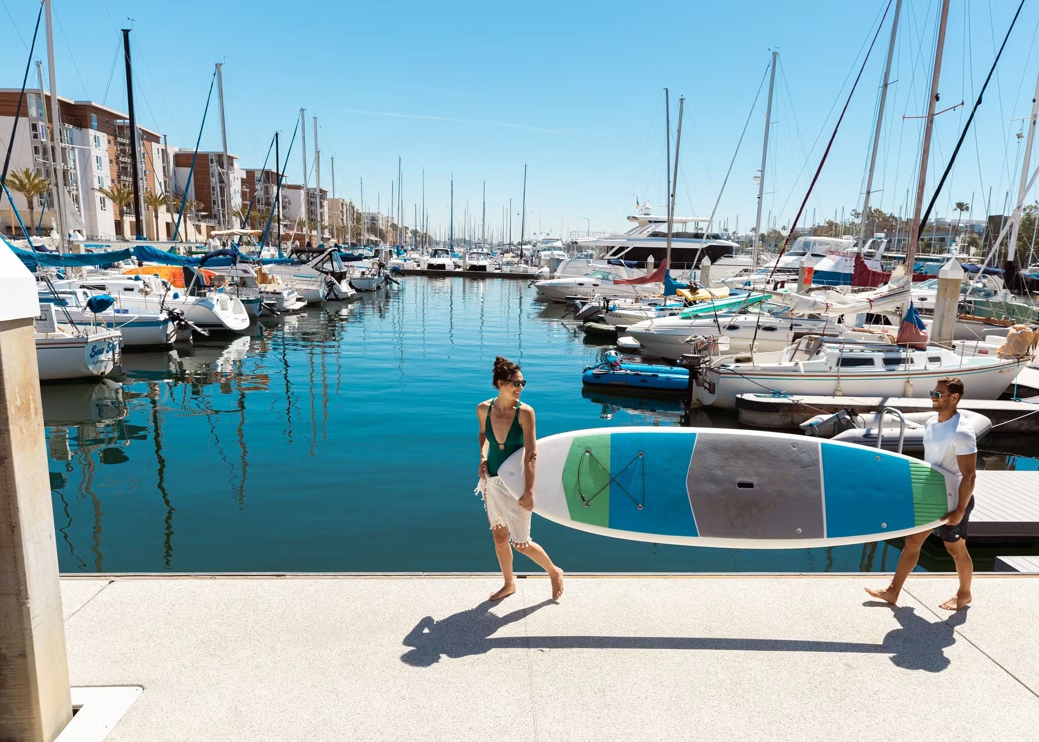 A marina with boats docked along the water's edge, a clear sky, and a pedestrian walkway.
