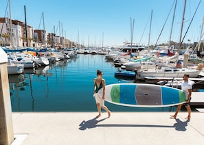 A marina with boats docked along the water's edge, a clear sky, and a pedestrian walkway.