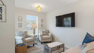 Un salon comprenant deux fauteuils, une table basse, un téléviseur fixé au mur et une fenêtre avec des stores.