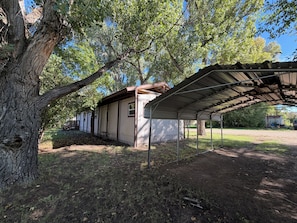 A covered outdoor area with a metal roof, surrounded by trees and a building with a white exterior.