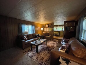 A cozy living room with a sofa, armchair, and coffee table. There is a kitchen area in the background.