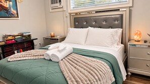 A neatly made bed with a green and beige duvet, a pair of rolled towels, and a Himalayan salt lamp on a bedside table.