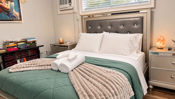 A neatly made bed with a green and beige duvet, a pair of rolled towels, and a Himalayan salt lamp on a bedside table.