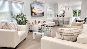 A modern living room with a glass coffee table, white sofas, and a kitchen area in the background.