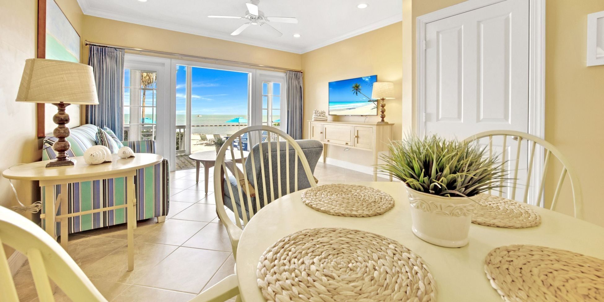 A room with a dining table, chairs, a ceiling fan, a television, and a view of the beach.
