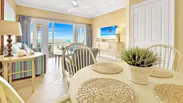 A room with a dining table, chairs, a ceiling fan, a television, and a view of the beach.