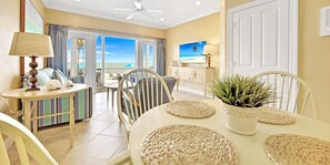 A room with a dining table, chairs, a ceiling fan, a television, and a view of the beach.