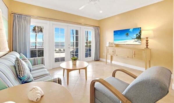 A room with a sofa, a round table, a chair, a television, and a view of the beach through the sliding doors.