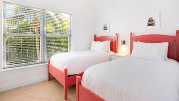A room with two beds, white bedding, a red headboard, a window with blinds, and a wall-mounted light.