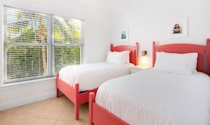 A room with two beds, white bedding, a red headboard, a window with blinds, and a wall-mounted light.