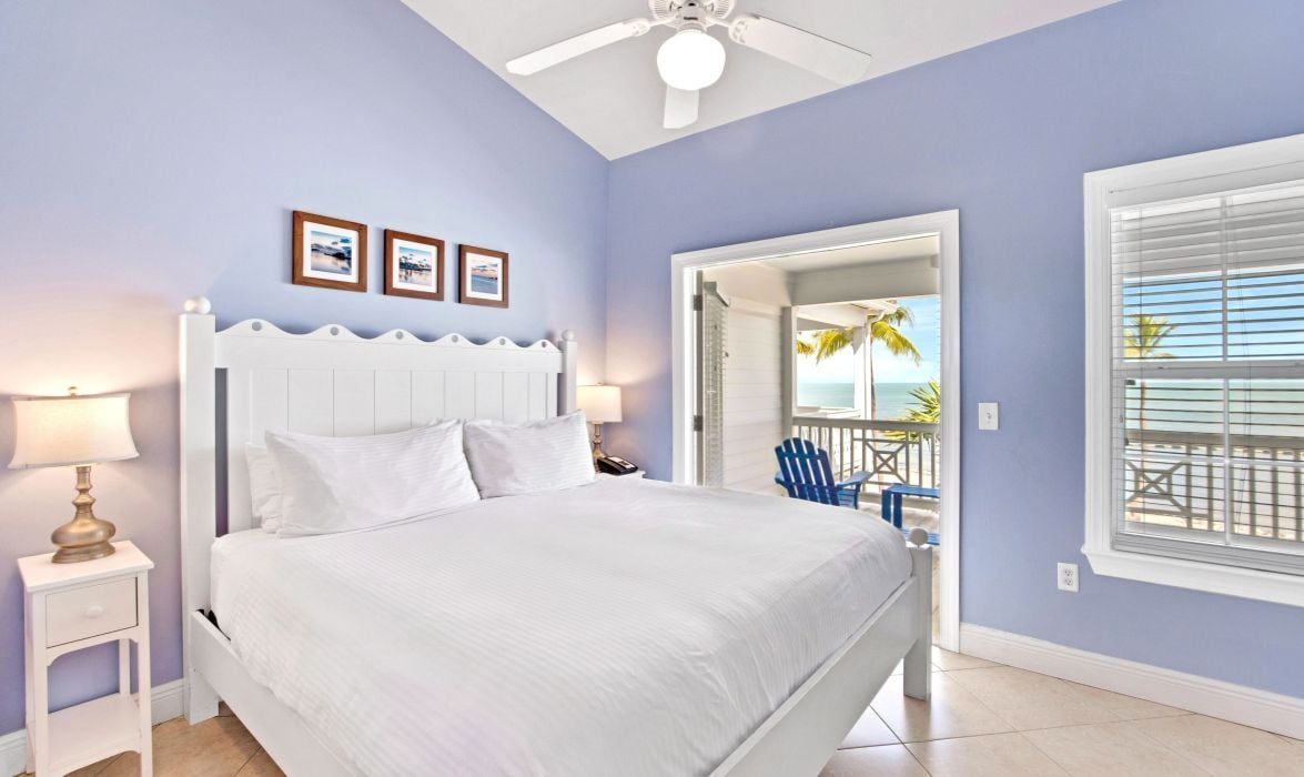 A bedroom with a white bed, a ceiling fan, and a view of the beach through a window.