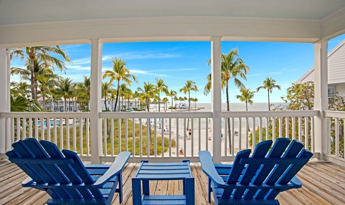 A covered outdoor area with a wooden deck, blue Adirondack chairs, and a view of palm trees and a beach.