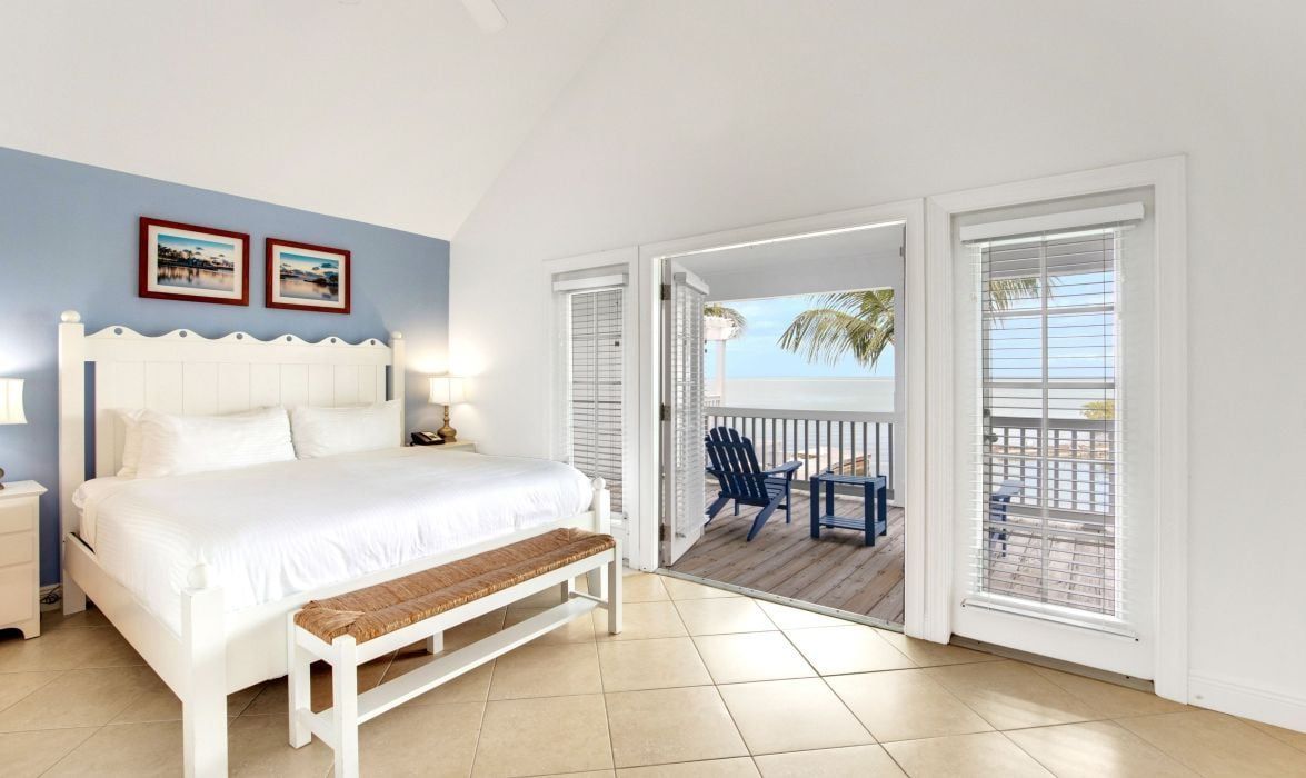 A bedroom with a bed, bedside table, bench, and a view of a balcony with chairs.