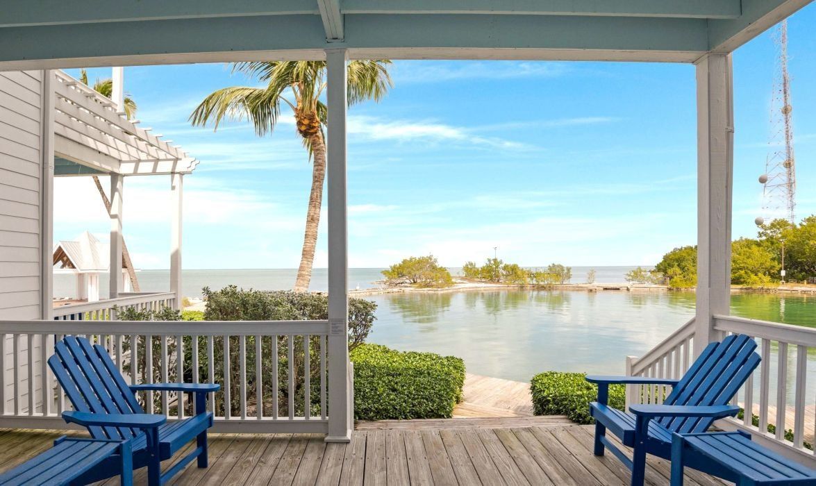 A covered porch with two blue Adirondack chairs, a view of a calm body of water, and a clear sky.