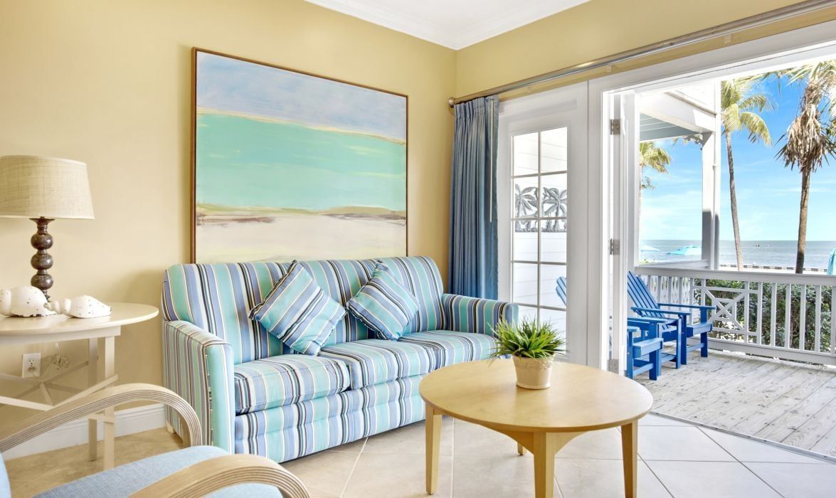 A room with a striped sofa, a round table, a lamp, and a view of the beach through a glass door.