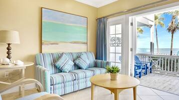 A room with a striped sofa, a round table, a lamp, and a view of the beach through a glass door.