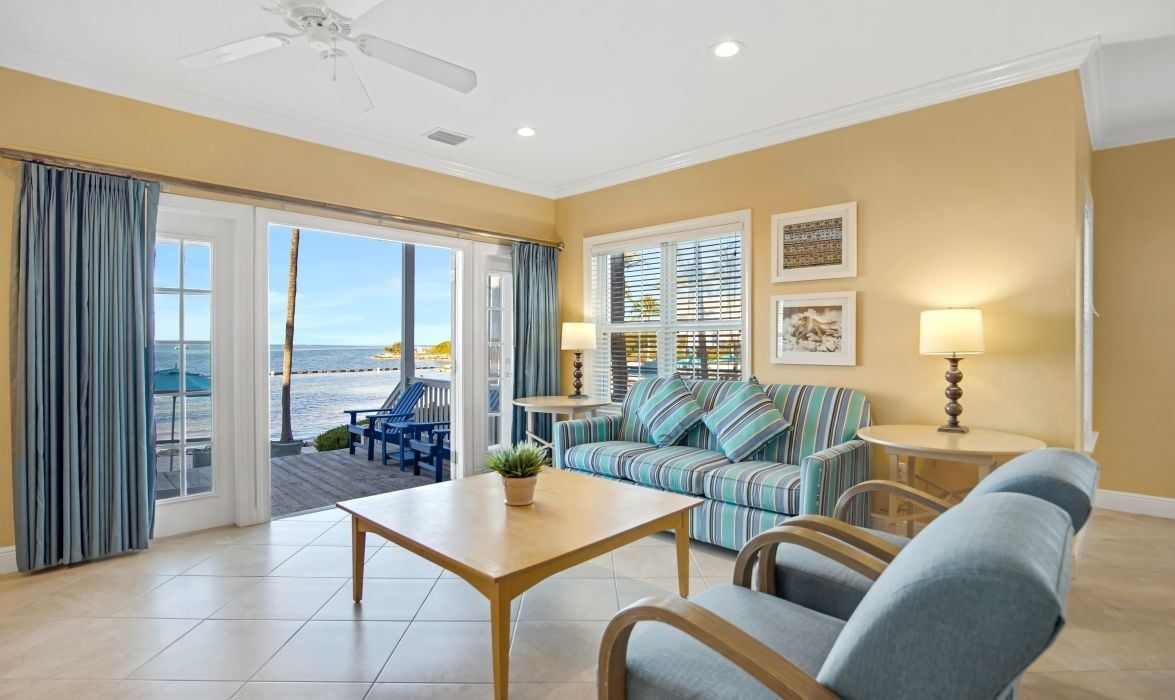 A room with a view of the sea, a sofa, a chair, a table, and a lamp.