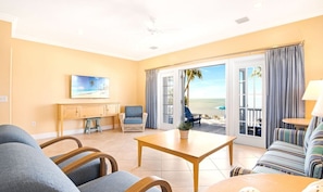 A room with a large sliding glass door leading to a balcony with a view of the ocean. There is a wooden table, a sofa, and a chair with cushions.