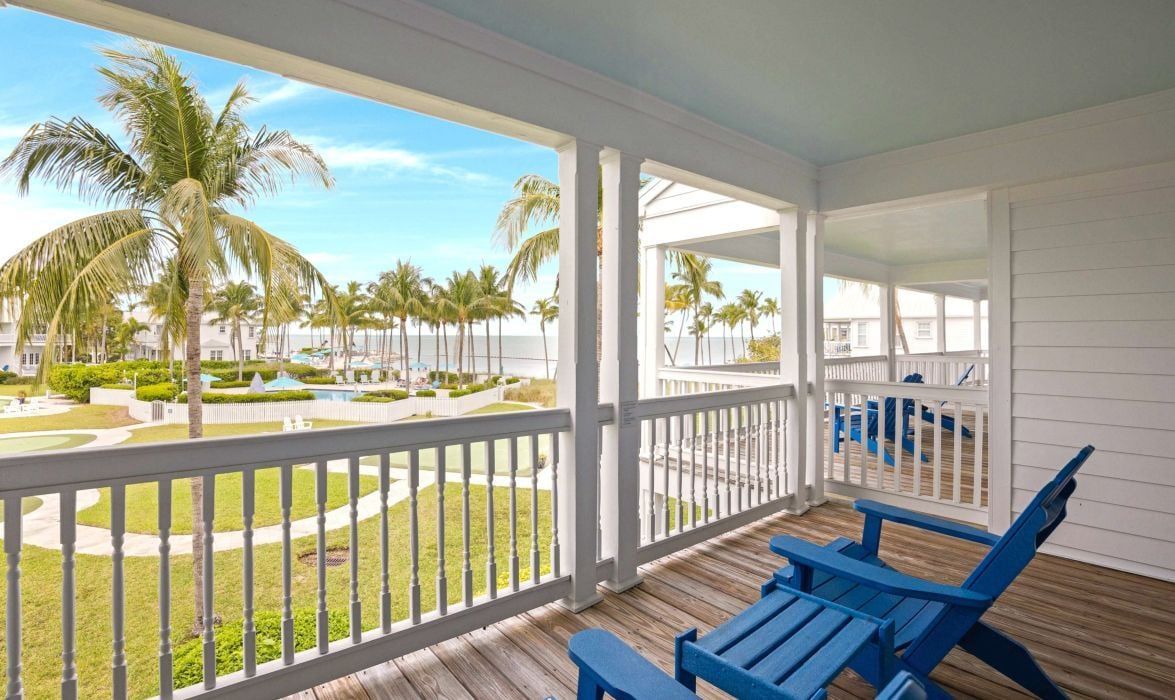 A covered outdoor area with a wooden deck, blue chairs, and a view of a beach with palm trees.