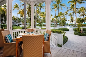 A covered outdoor seating area with wicker furniture and a table set for two, overlooking a landscaped garden with palm trees.