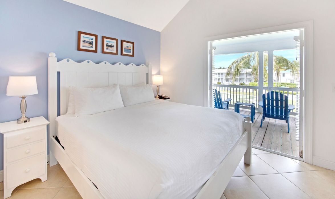 A hotel room with a large bed, a nightstand with a lamp, and a view of the outdoor balcony.