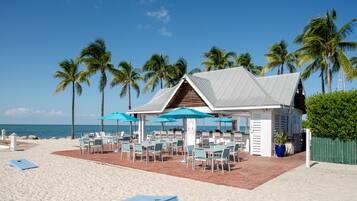 Un restaurant situé en bord de mer, avec une terrasse extérieure, un patio couvert et des palmiers en arrière-plan.