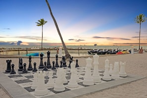 A chessboard set up on a beach with palm trees and a sunset in the background.